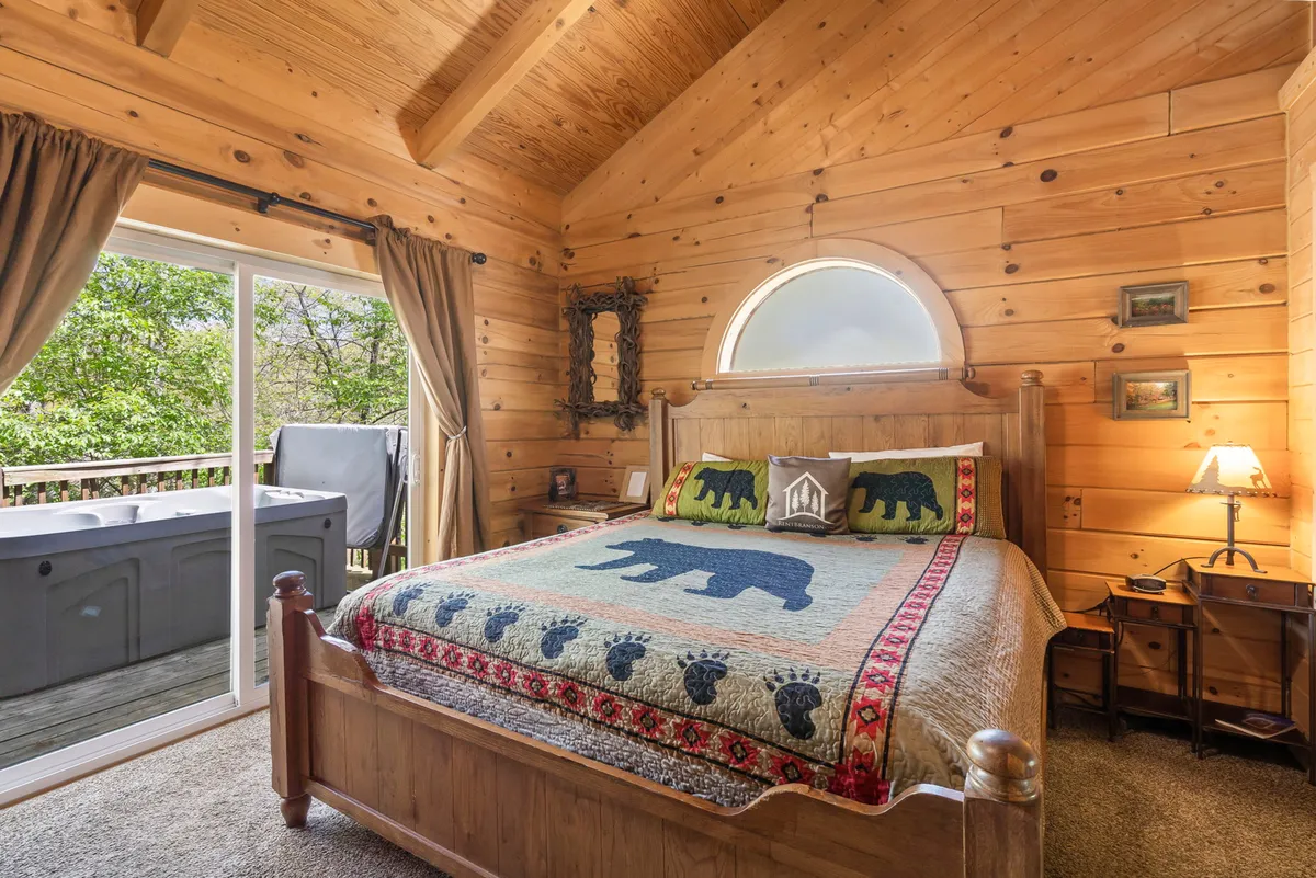 A glimpse into a rustic cabin bedroom with a bear-themed quilt, wood-paneled walls, and a large window offering views of the hot tub just outside. The space feels warm and inviting, seamlessly blending indoor comfort with outdoor romance.
