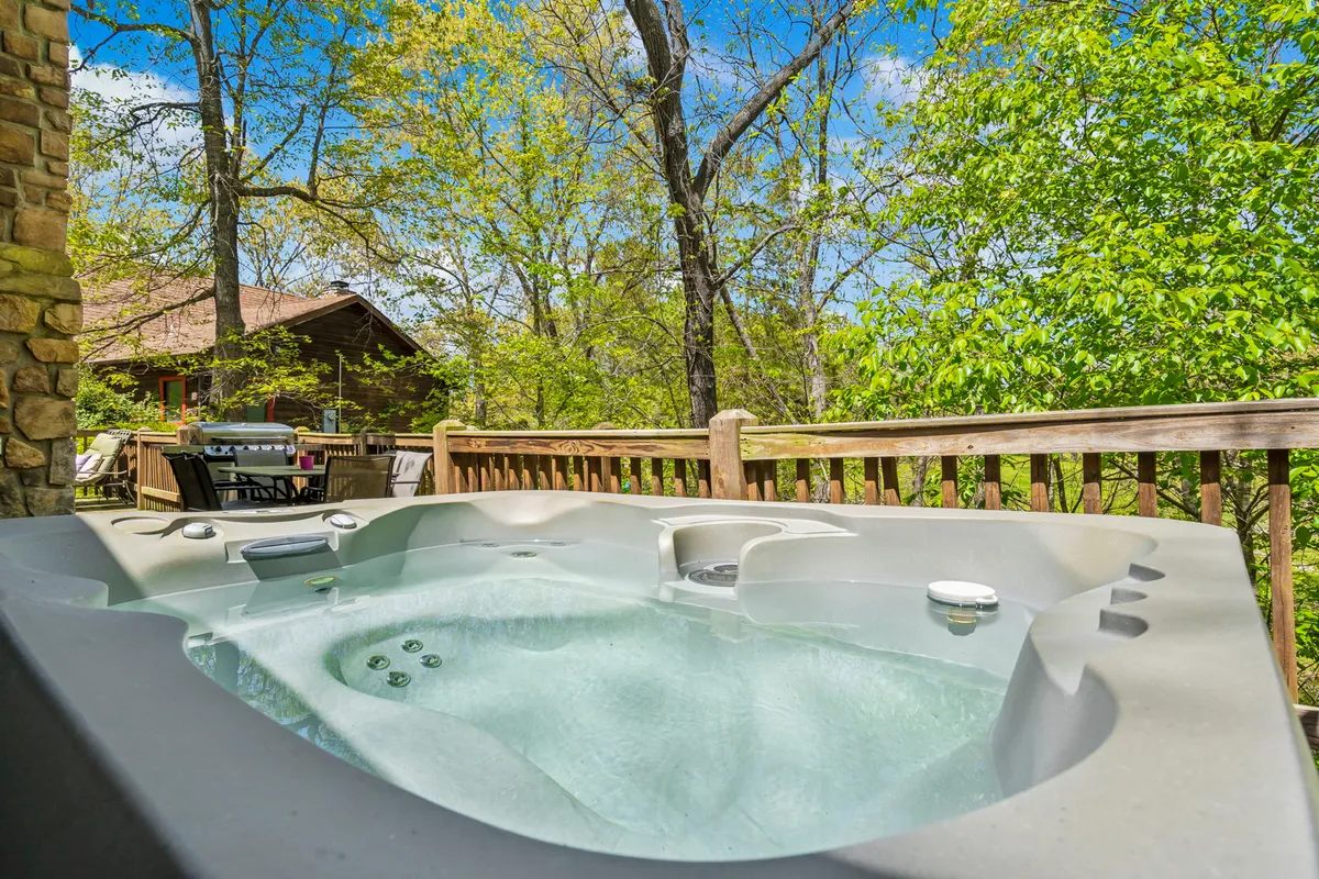A tranquil view of a bubbling hot tub tucked on a wooden deck, framed by tree branches and a sturdy deck railing. The surrounding woods offer privacy and a sense of immersion in nature, while the hot tub promises relaxation under the open sky.