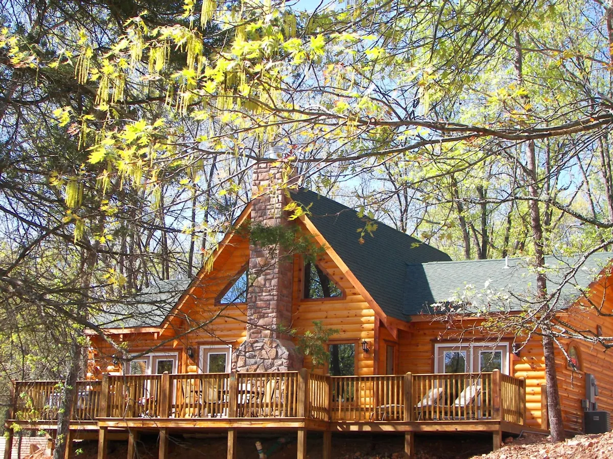 A cozy log cabin stands amidst lush greenery, featuring a prominent stone chimney and an inviting wooden deck. The setting evokes tranquility and seclusion, ideal for a romantic retreat. The deck sets the stage for relaxing activities like candlelit dinners or stargazing.