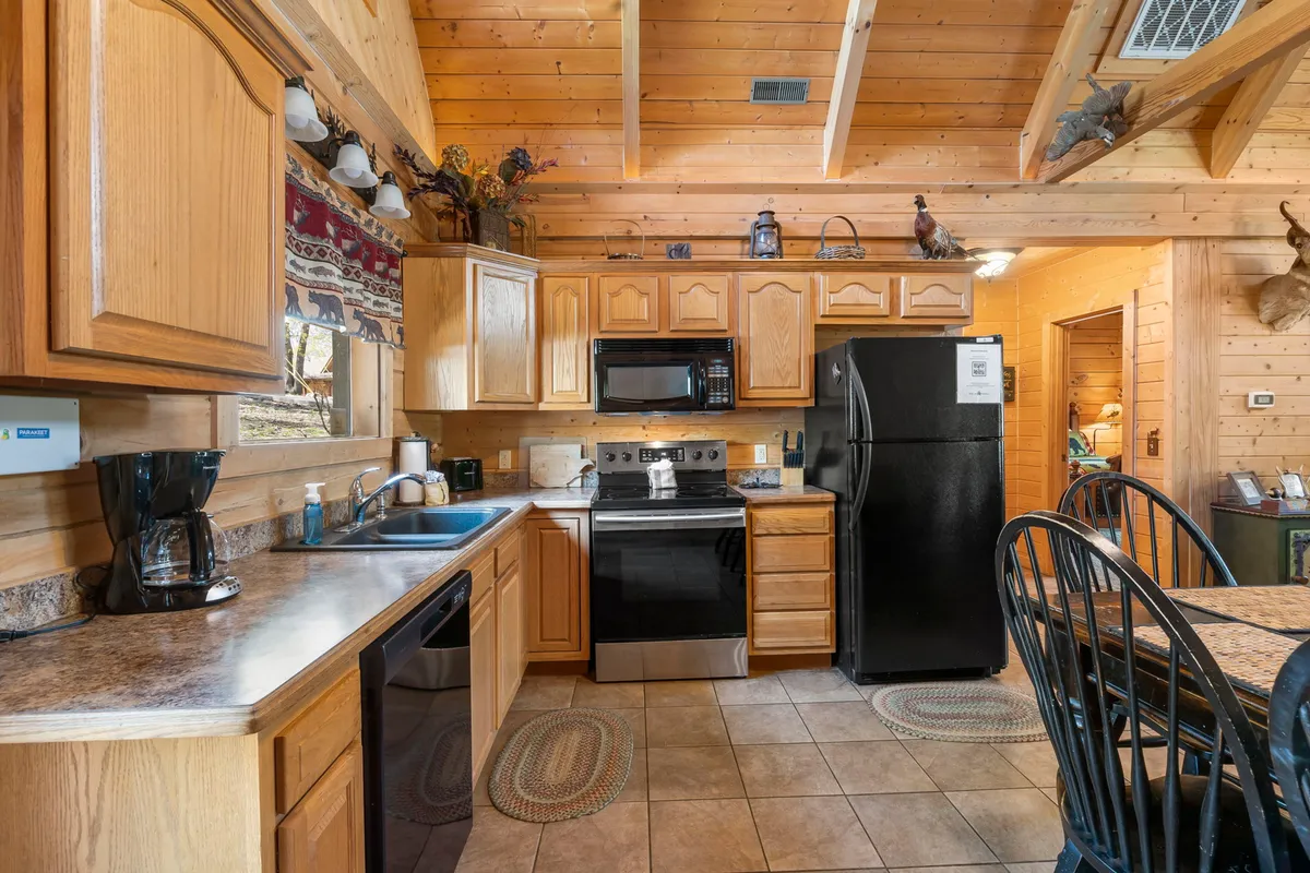 A warm, inviting kitchen scene with a prominent kitchen island, a spacious wooden dining table, sleek black appliances, and wooden cabinetry. Sunlight streams in through a window, illuminating the prep spaces and dining area.