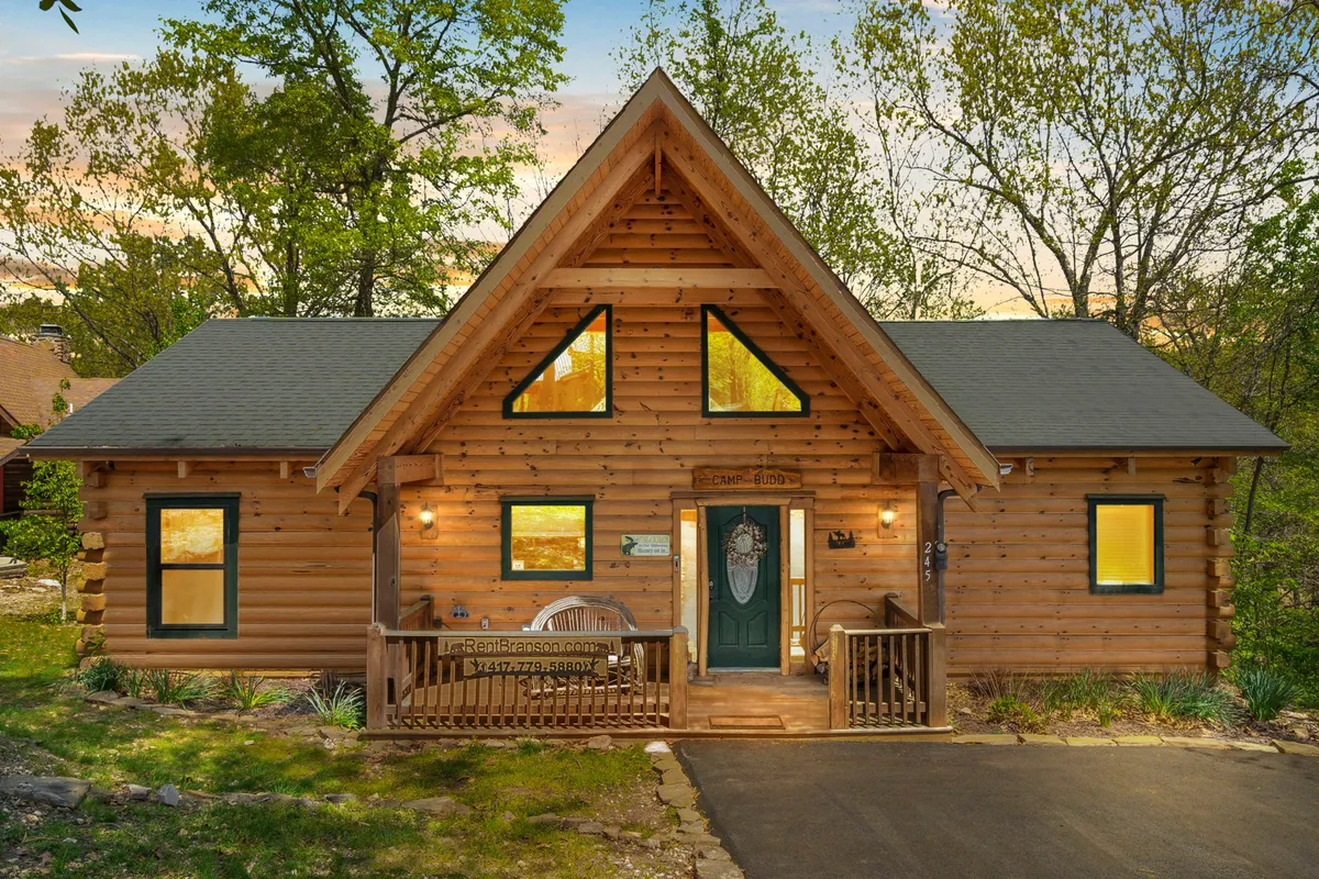 The image features a charming log cabin nestled among the trees, with large front windows, a decorative wooden front door, and a welcoming porch complete with cozy outdoor seating. The tranquil forest setting and inviting porch evoke a sense of peace, perfect for relaxing or gathering with loved ones. Warm evening light bathes the cabin, hinting at sunset views and memorable outdoor moments.