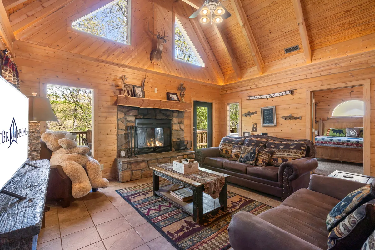 A spacious great room featuring a majestic stone fireplace, lofty wooden beams, large windows that let in abundant natural light, a plush sofa, and a central coffee table. The setting is cozy yet grand, seamlessly blending rustic charm with modern relaxation and inviting areas for gathering.