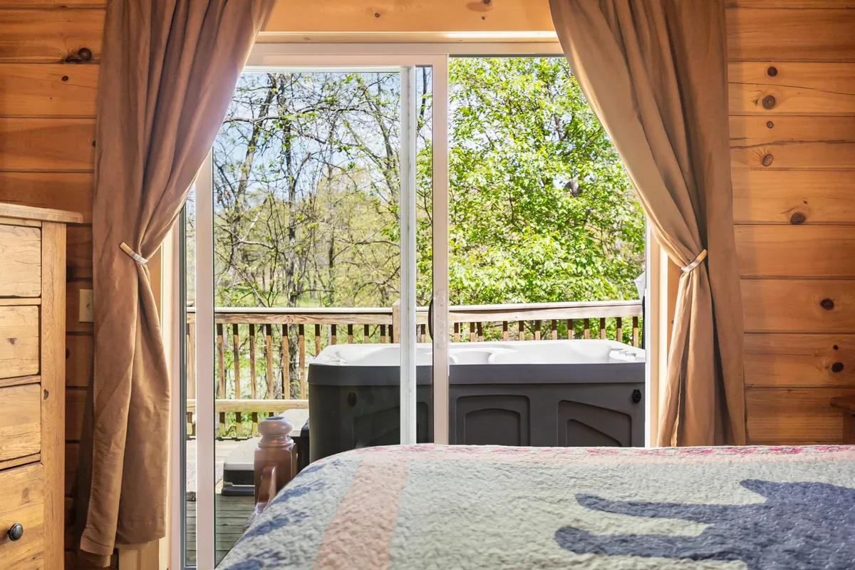 A cozy bedroom with a stylish quilt on the bed, sliding glass doors leading to a private deck, and a hot tub just outside. The room is surrounded by natural greenery, creating a seamless connection between interior comfort and the peaceful woods outside.