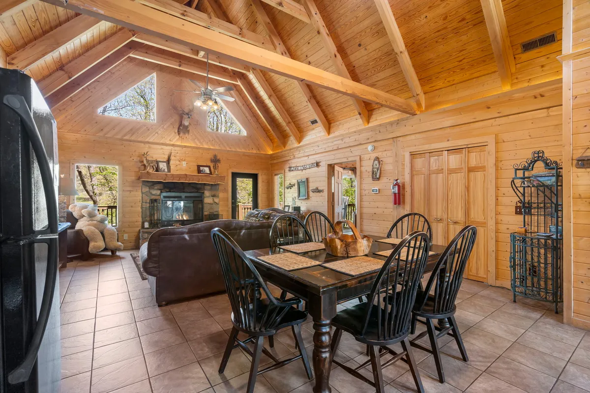 A cozy dining area featuring a large wooden dining table, rustic wooden beams, a stone fireplace, and expansive windows offering scenic views. The space is set for gatherings, highlighting the communal aspect of shared meals and group activities.