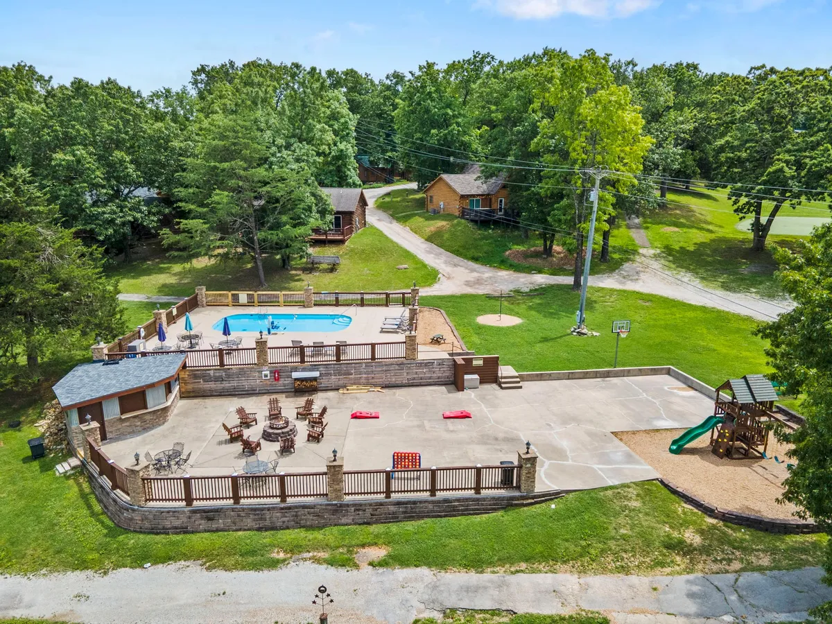 This image features a vibrant family recreation area with a swimming pool, patio, playground, and basketball hoop, all surrounded by green lawns and trees. The scene shows different activity zones in one view, capturing both the pool and active play spaces.