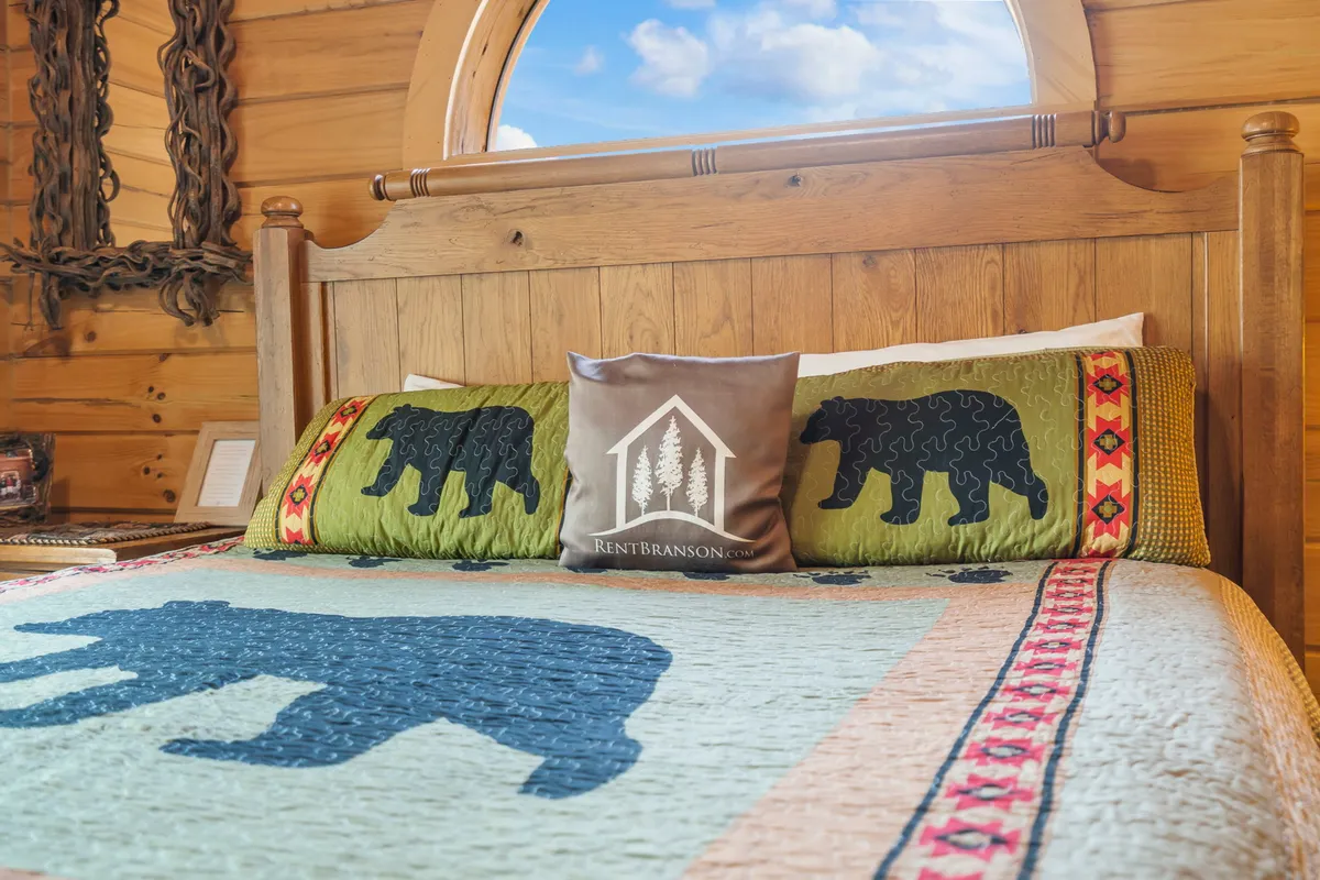 A welcoming queen bedroom with a wooden bed frame, bear-themed quilt, and a circular window letting in soft natural light. The decor is cozy and rustic, echoing the tranquility of the surrounding woods.