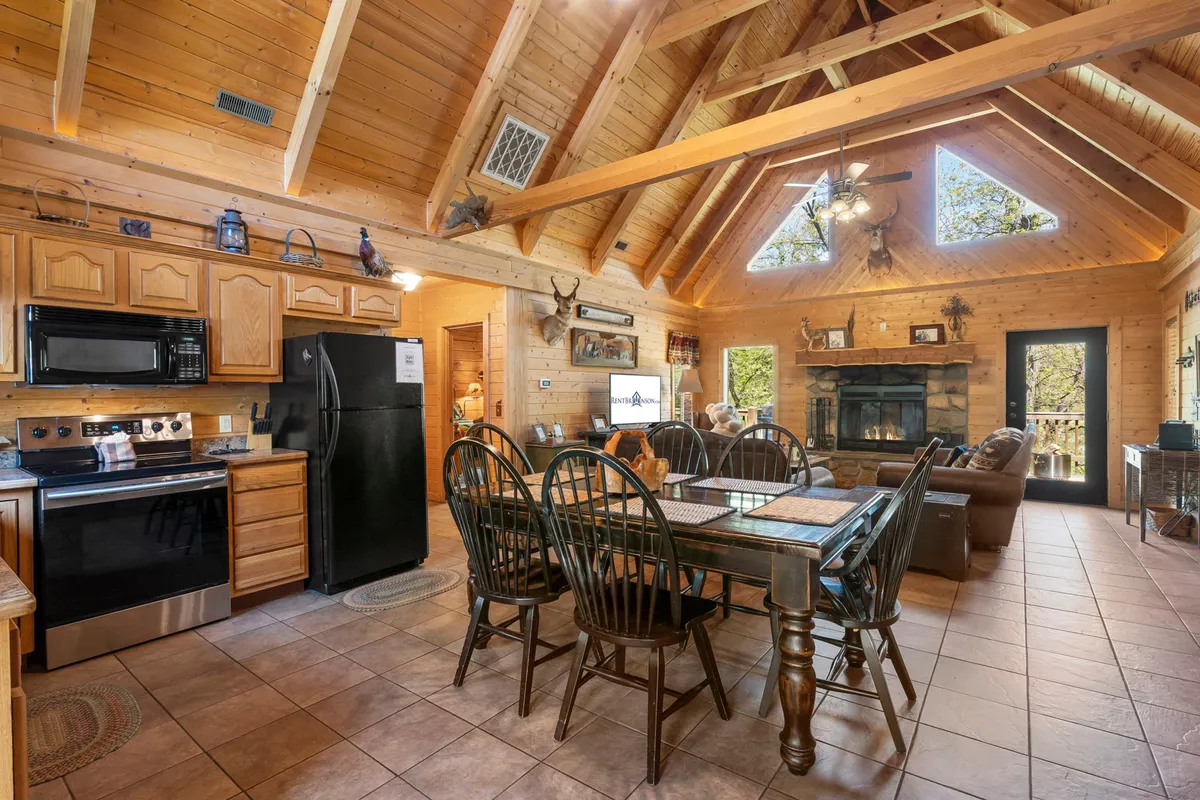 An angle from the dining area, capturing a large wooden dining table set before a stone fireplace, with vaulted timbered ceilings overhead and the kitchen visible in the background. The room feels spacious and welcoming, highlighting the seamless flow between dining, cooking, and relaxing.
